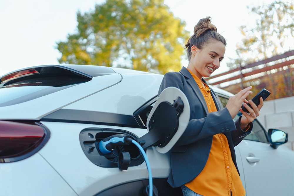 Woman leaning against her electric vehicle while it charges; she's using her mobile phone and smiling.