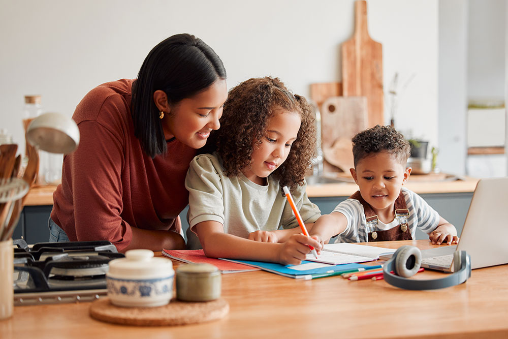 Mom helping daughter with homework at kitchen counter. Young son participating.