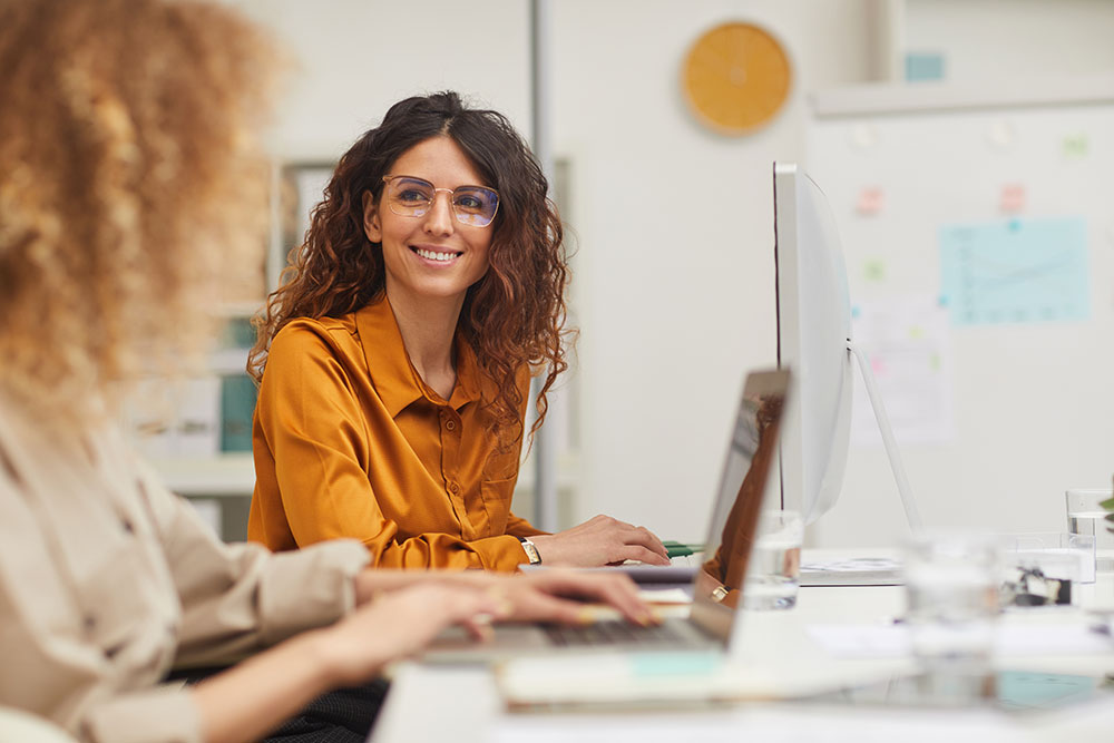 Woman working at laptop and smiling at co-worker in office.